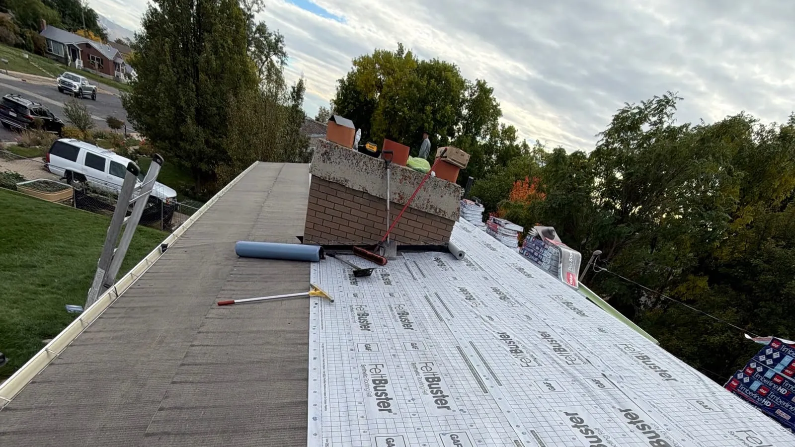 New synthetic underlayment being rolled out on a roof beside a brick chimney, with shingle bundles staged nearby